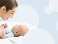 Uśmiechnięta mama pochyla się nad dzieckiem, które leży na białym tle. To magiczny moment, kiedy dziecko zaczyna mówić mama.