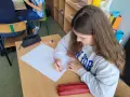 Dziewczynka rysuje w zeszycie, wykonując zadania z polskiego klasa 3. Na biurku leży czerwony piórnik.