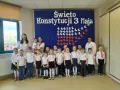 Dzieci w odświętnych strojach pozują do zdjęcia z nauczycielkami na tle niebieskiej kotary z napisem "Święto Konstytucji 3 Maja".