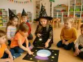 Dzieci w czarodziejskich czapkach na podłodze, przygotowują się do wróżb andrzejkowych w szkole. Jedna z dziewczynek świeci latarką na magiczną kulę.