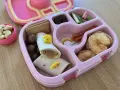 Różowy lunchbox z pysznym, zdrowym śniadaniem do szkoły: wrap z kurczakiem, suszone jabłka, kiwi, orzechy i czekoladowe kuleczki.