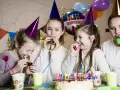 Radosne dzieci w czapkach urodzinowych dmuchają w gwizdki, świętując w domu. W tle balony i tort. Idealne animacje na urodziny w domu!