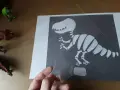 Ręka trzyma przezroczystą folię nad rysunkiem szkieletu dinozaura. To ciekawa szkielet dinozaura praca plastyczna.