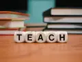Kostki z literami tworzą słowo "TEACH". W tle stosy książek, idealne do nauki wyrazów na b dla dzieci.