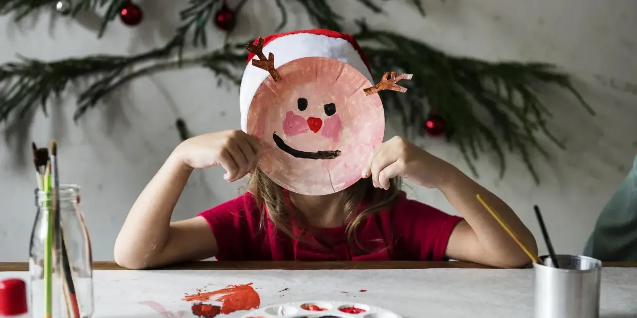 Dziecko w czapce Mikołaja tworzy świąteczne prace plastyczne w przedszkolu, trzymając przed twarzą własnoręcznie zrobioną maskę renifera.