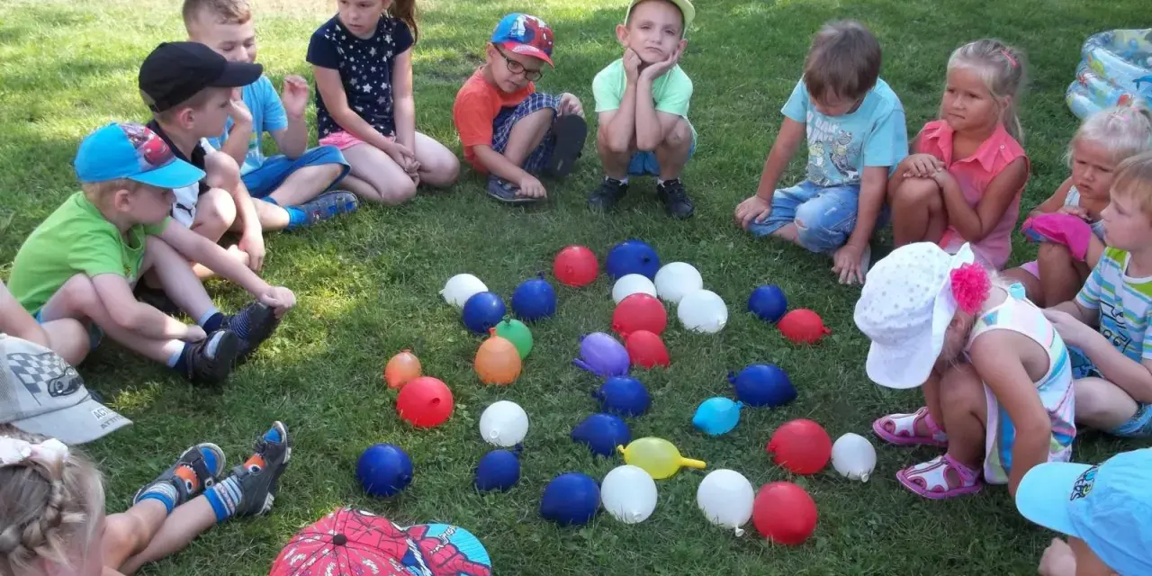 Dzieci siedzą na trawie, otoczone kolorowymi balonami. To wesołe zabawy z balonami dla dzieci na świeżym powietrzu.