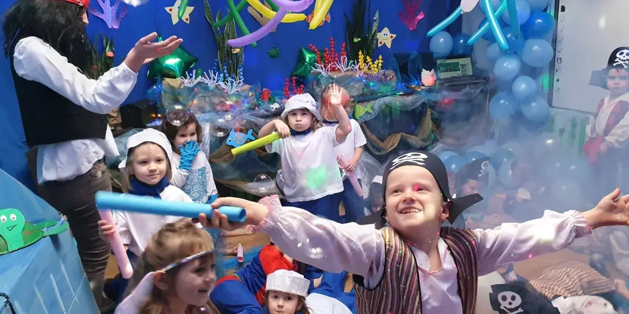 Dzieci w strojach piratów i wróżek bawią się na podwodnym balu karnawałowym. Balony i dekoracje tworzą atmosferę morskiej przygody.