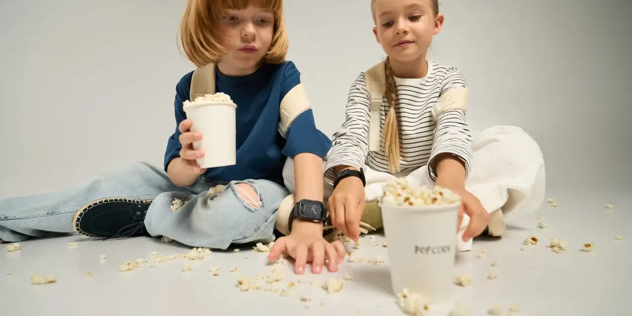 Dzieci siedzą na podłodze, jedząc popcorn z kubków. To idealny moment na wspólne oglądanie bajek, a kieszonkowe dla dziecka na przekąski zawsze się przyda.