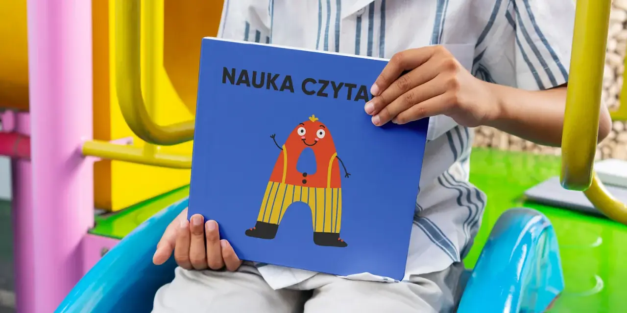 Chłopiec trzyma książkę "Nauka czytania dla dzieci 6 lat" z zabawną literką "A" na okładce.