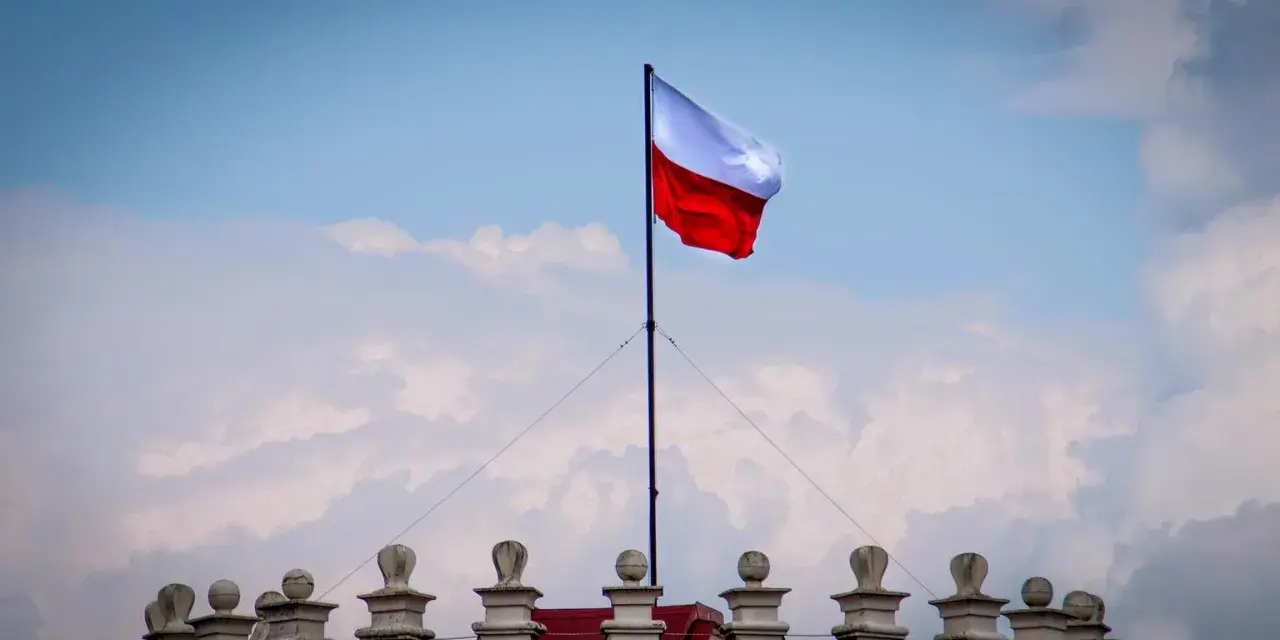 Biało-czerwona flaga powiewa na maszcie nad zabytkowym budynkiem. Wiatr rozwiewa ją, jakby niosła słowa o Polsce.