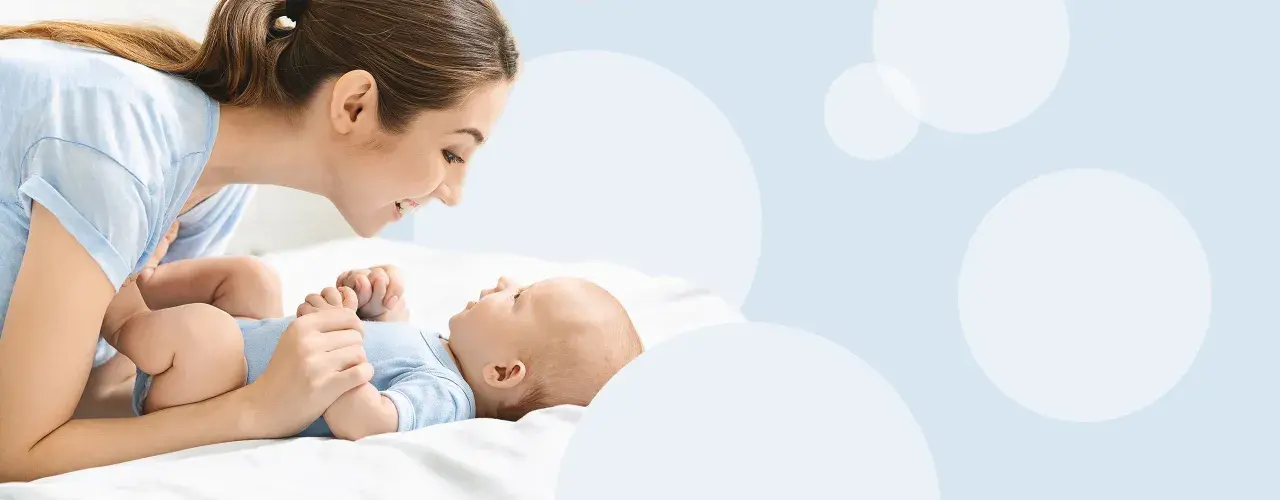 Uśmiechnięta mama pochyla się nad dzieckiem, które leży na białym tle. To magiczny moment, kiedy dziecko zaczyna mówić mama.
