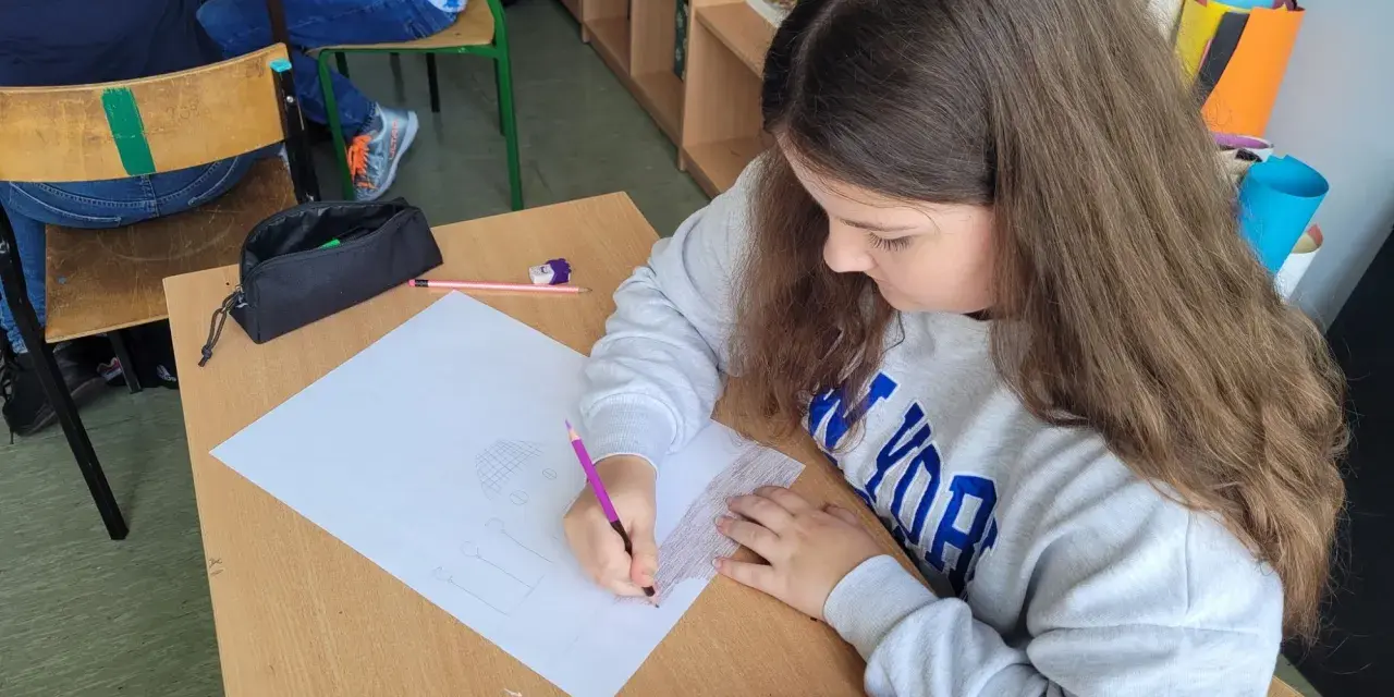 Dziewczynka rysuje w zeszycie, wykonując zadania z polskiego klasa 3. Na biurku leży czerwony piórnik.