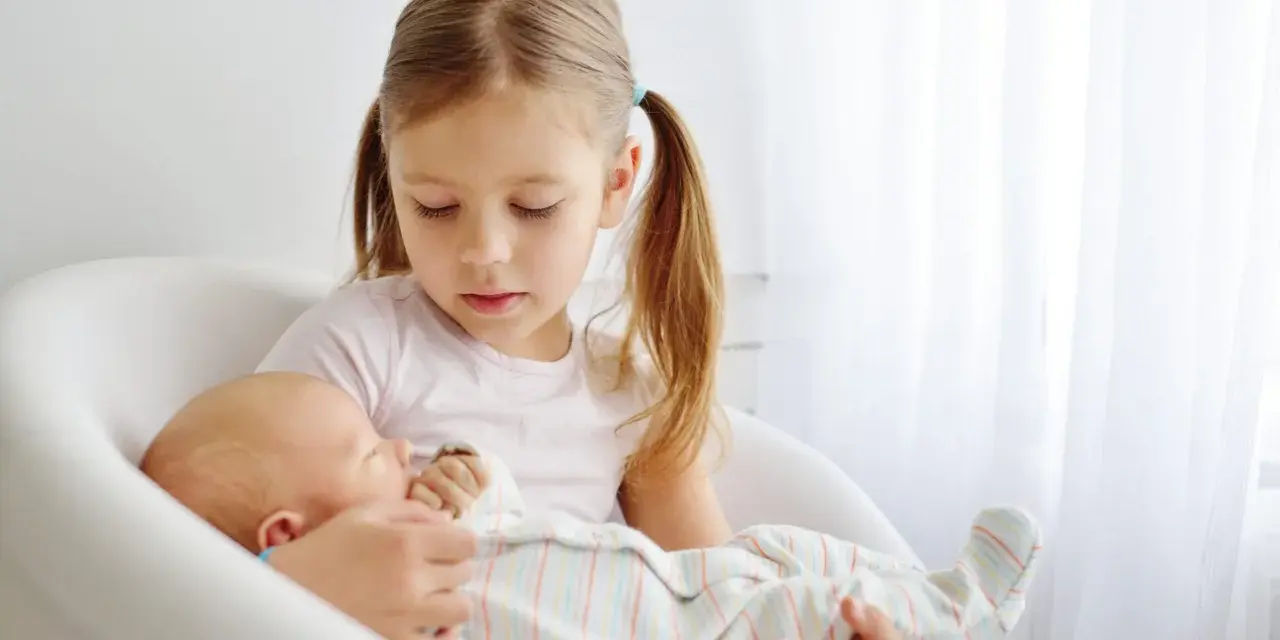 Starsza siostra delikatnie trzyma noworodka, ucząc się jak przygotować dziecko na rodzeństwo.