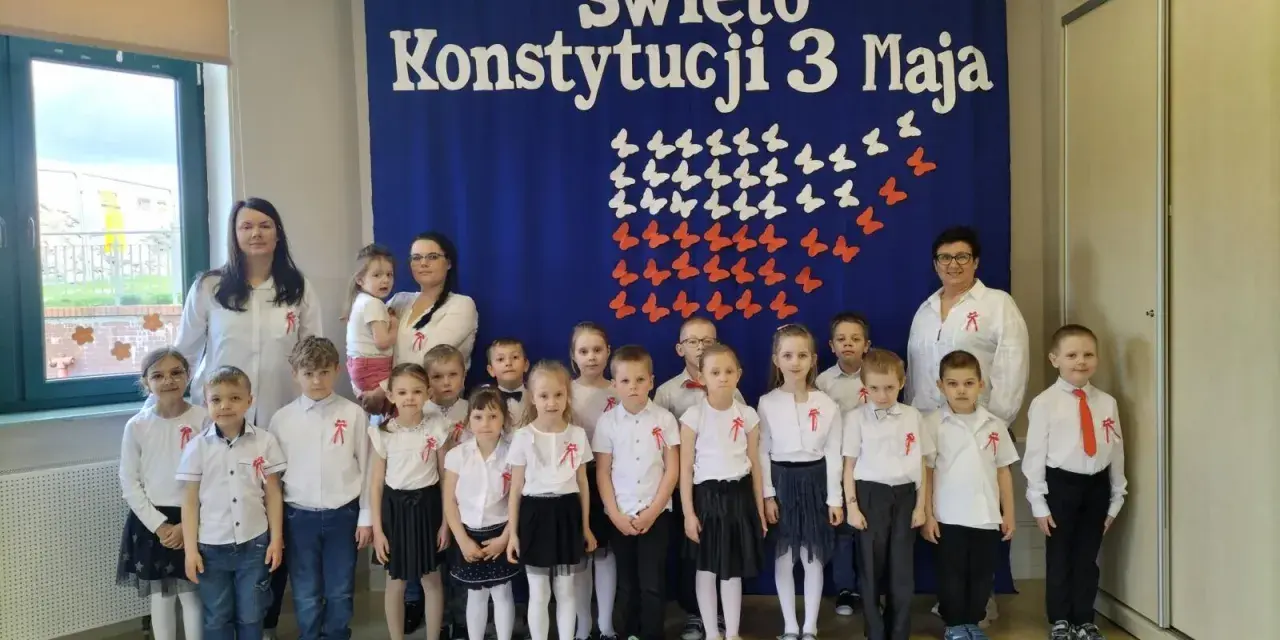 Dzieci w odświętnych strojach pozują do zdjęcia z nauczycielkami na tle niebieskiej kotary z napisem "Święto Konstytucji 3 Maja".