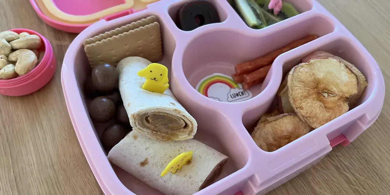Różowy lunchbox z pysznym, zdrowym śniadaniem do szkoły: wrap z kurczakiem, suszone jabłka, kiwi, orzechy i czekoladowe kuleczki.