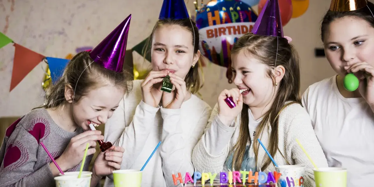 Radosne dzieci w czapkach urodzinowych dmuchają w gwizdki, świętując w domu. W tle balony i tort. Idealne animacje na urodziny w domu!