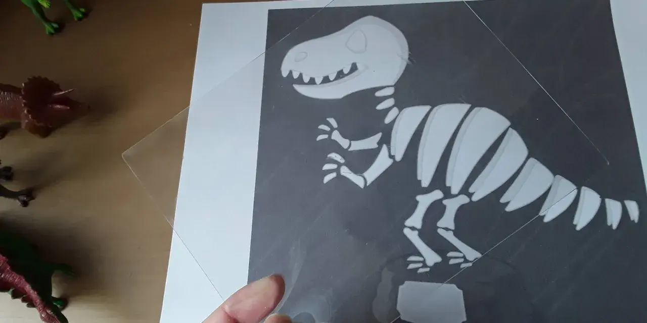 Ręka trzyma przezroczystą folię nad rysunkiem szkieletu dinozaura. To ciekawa szkielet dinozaura praca plastyczna.
