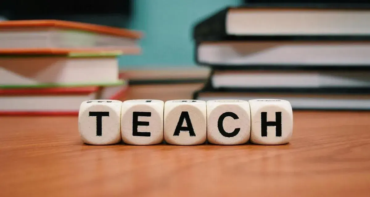 Kostki z literami tworzą słowo "TEACH". W tle stosy książek, idealne do nauki wyrazów na b dla dzieci.