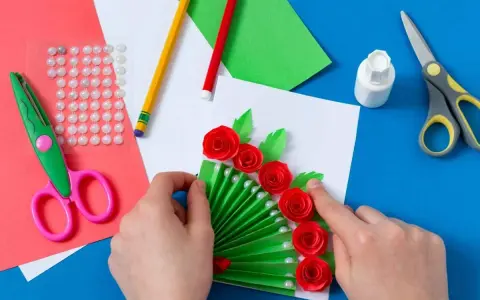Ręcznie robiona laurka dla mamy z zielonym wachlarzem, czerwonymi różami i perłami. Wokół materiały do tworzenia: nożyczki, klej, kredki, naklejki.