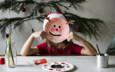 Dziecko w czapce Mikołaja tworzy świąteczne prace plastyczne w przedszkolu, trzymając przed twarzą własnoręcznie zrobioną maskę renifera.