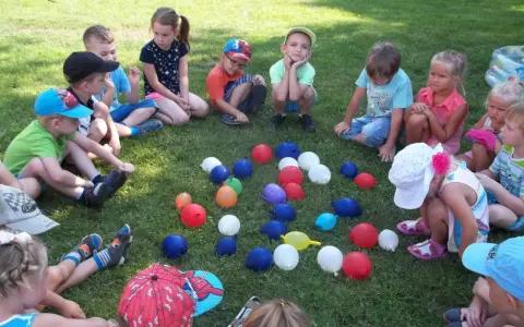 Dzieci siedzą na trawie, otoczone kolorowymi balonami. To wesołe zabawy z balonami dla dzieci na świeżym powietrzu.