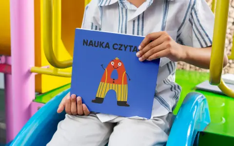 Chłopiec trzyma książkę "Nauka czytania dla dzieci 6 lat" z zabawną literką "A" na okładce.