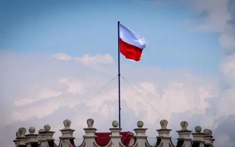 Biało-czerwona flaga powiewa na maszcie nad zabytkowym budynkiem. Wiatr rozwiewa ją, jakby niosła słowa o Polsce.