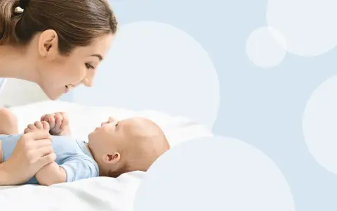 Uśmiechnięta mama pochyla się nad dzieckiem, które leży na białym tle. To magiczny moment, kiedy dziecko zaczyna mówić mama.