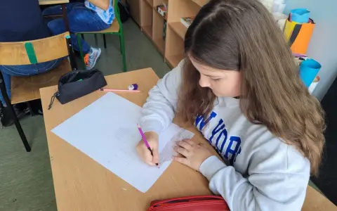Dziewczynka rysuje w zeszycie, wykonując zadania z polskiego klasa 3. Na biurku leży czerwony piórnik.