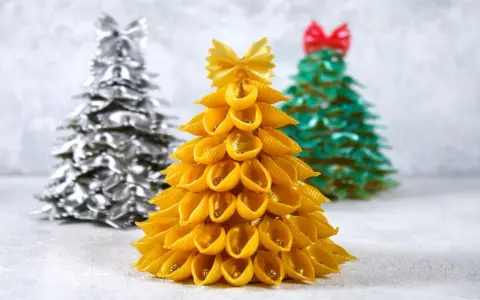 Choinka DIY dla dzieci z makaronu muszelki, ozdobiona złotym brokatem. W tle choinki z makaronu farfalle w kolorze srebrnym i zielonym.