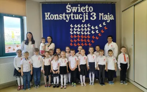 Dzieci w odświętnych strojach pozują do zdjęcia z nauczycielkami na tle niebieskiej kotary z napisem "Święto Konstytucji 3 Maja".