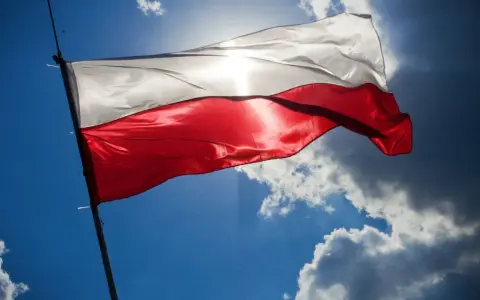Biało-czerwona flaga powiewa na tle błękitnego nieba z chmurami. Wiatr rozwiewa ją, przypominając o pięknych cytatach o ojczyźnie.
