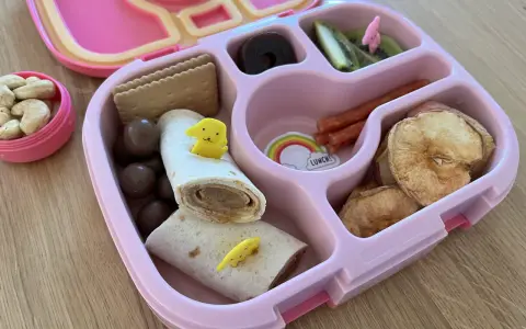 Różowy lunchbox z pysznym, zdrowym śniadaniem do szkoły: wrap z kurczakiem, suszone jabłka, kiwi, orzechy i czekoladowe kuleczki.