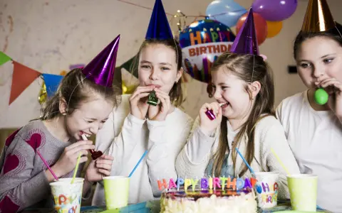 Radosne dzieci w czapkach urodzinowych dmuchają w gwizdki, świętując w domu. W tle balony i tort. Idealne animacje na urodziny w domu!