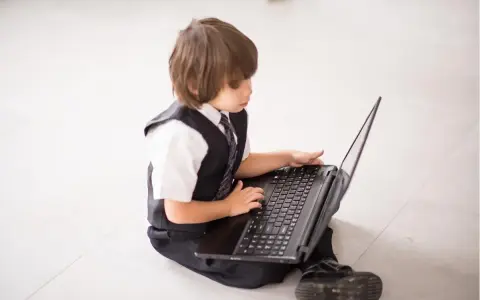Elegancki chłopiec w garniturze odkrywa świat w internecie, siedząc na podłodze z laptopem.