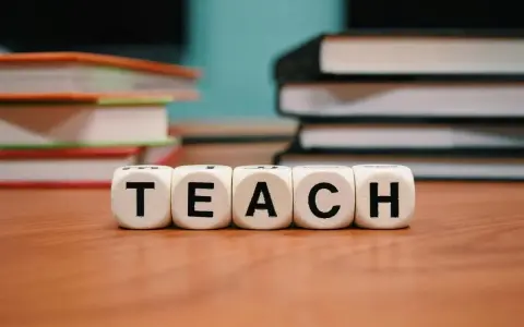 Kostki z literami tworzą słowo "TEACH". W tle stosy książek, idealne do nauki wyrazów na b dla dzieci.