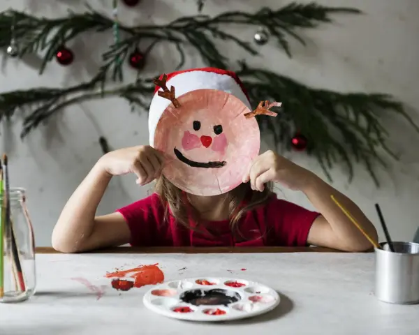 Dziecko w czapce Mikołaja tworzy świąteczne prace plastyczne w przedszkolu, trzymając przed twarzą własnoręcznie zrobioną maskę renifera.
