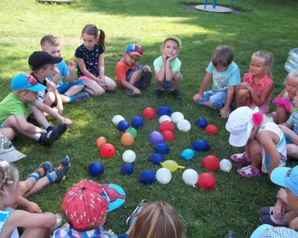 Dzieci siedzą na trawie, otoczone kolorowymi balonami. To wesołe zabawy z balonami dla dzieci na świeżym powietrzu.