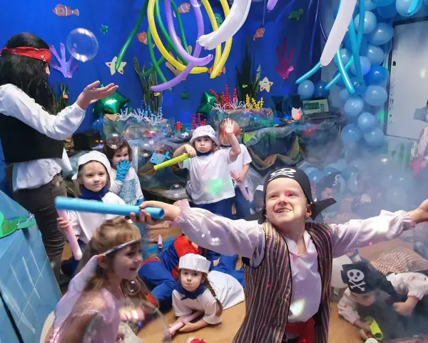 Dzieci w strojach piratów i wróżek bawią się na podwodnym balu karnawałowym. Balony i dekoracje tworzą atmosferę morskiej przygody.