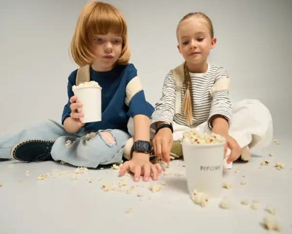Dzieci siedzą na podłodze, jedząc popcorn z kubków. To idealny moment na wspólne oglądanie bajek, a kieszonkowe dla dziecka na przekąski zawsze się przyda.