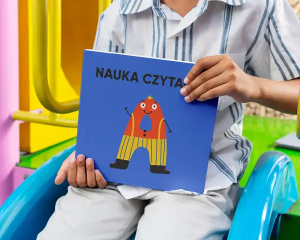 Chłopiec trzyma książkę "Nauka czytania dla dzieci 6 lat" z zabawną literką "A" na okładce.