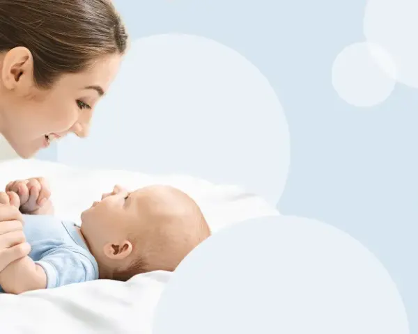 Uśmiechnięta mama pochyla się nad dzieckiem, które leży na białym tle. To magiczny moment, kiedy dziecko zaczyna mówić mama.