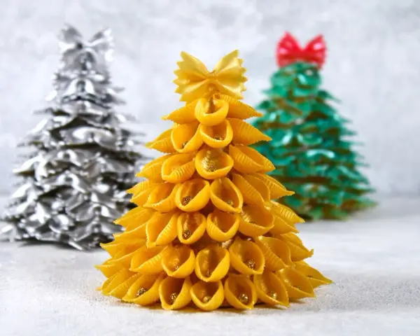 Choinka DIY dla dzieci z makaronu muszelki, ozdobiona złotym brokatem. W tle choinki z makaronu farfalle w kolorze srebrnym i zielonym.