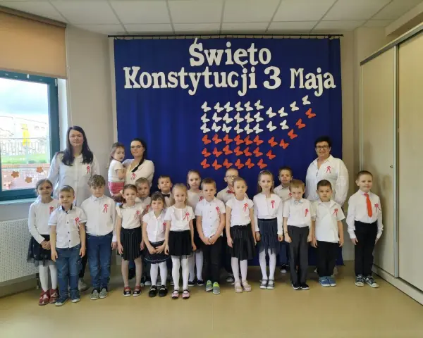 Dzieci w odświętnych strojach pozują do zdjęcia z nauczycielkami na tle niebieskiej kotary z napisem "Święto Konstytucji 3 Maja".