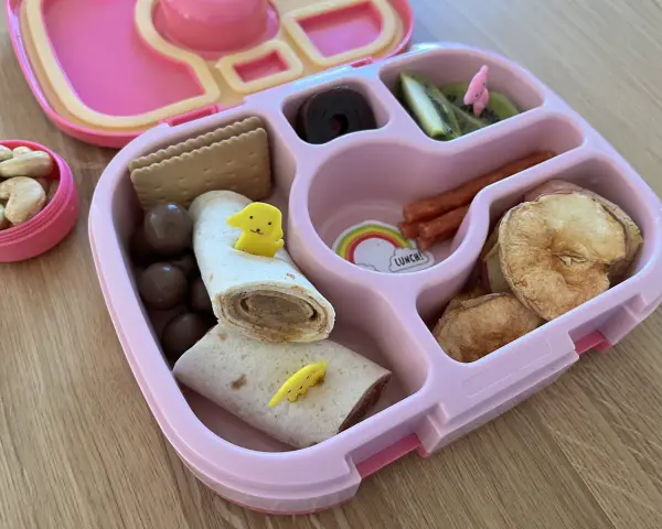 Różowy lunchbox z pysznym, zdrowym śniadaniem do szkoły: wrap z kurczakiem, suszone jabłka, kiwi, orzechy i czekoladowe kuleczki.