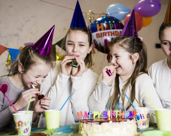 Radosne dzieci w czapkach urodzinowych dmuchają w gwizdki, świętując w domu. W tle balony i tort. Idealne animacje na urodziny w domu!