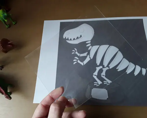 Ręka trzyma przezroczystą folię nad rysunkiem szkieletu dinozaura. To ciekawa szkielet dinozaura praca plastyczna.