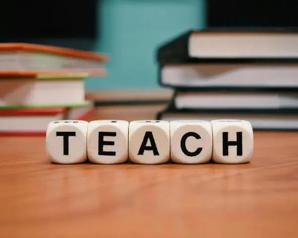 Kostki z literami tworzą słowo "TEACH". W tle stosy książek, idealne do nauki wyrazów na b dla dzieci.