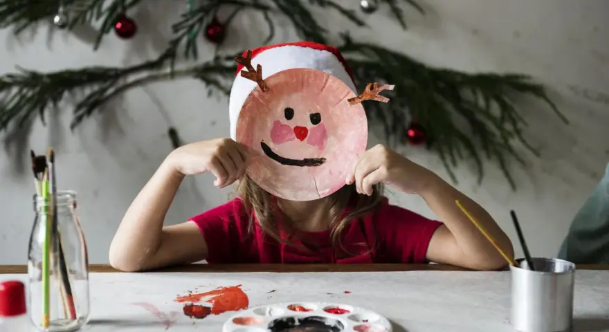 Dziecko w czapce Mikołaja tworzy świąteczne prace plastyczne w przedszkolu, trzymając przed twarzą własnoręcznie zrobioną maskę renifera.
