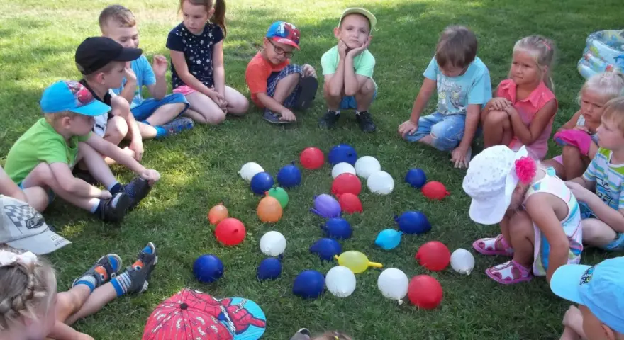 Dzieci siedzą na trawie, otoczone kolorowymi balonami. To wesołe zabawy z balonami dla dzieci na świeżym powietrzu.
